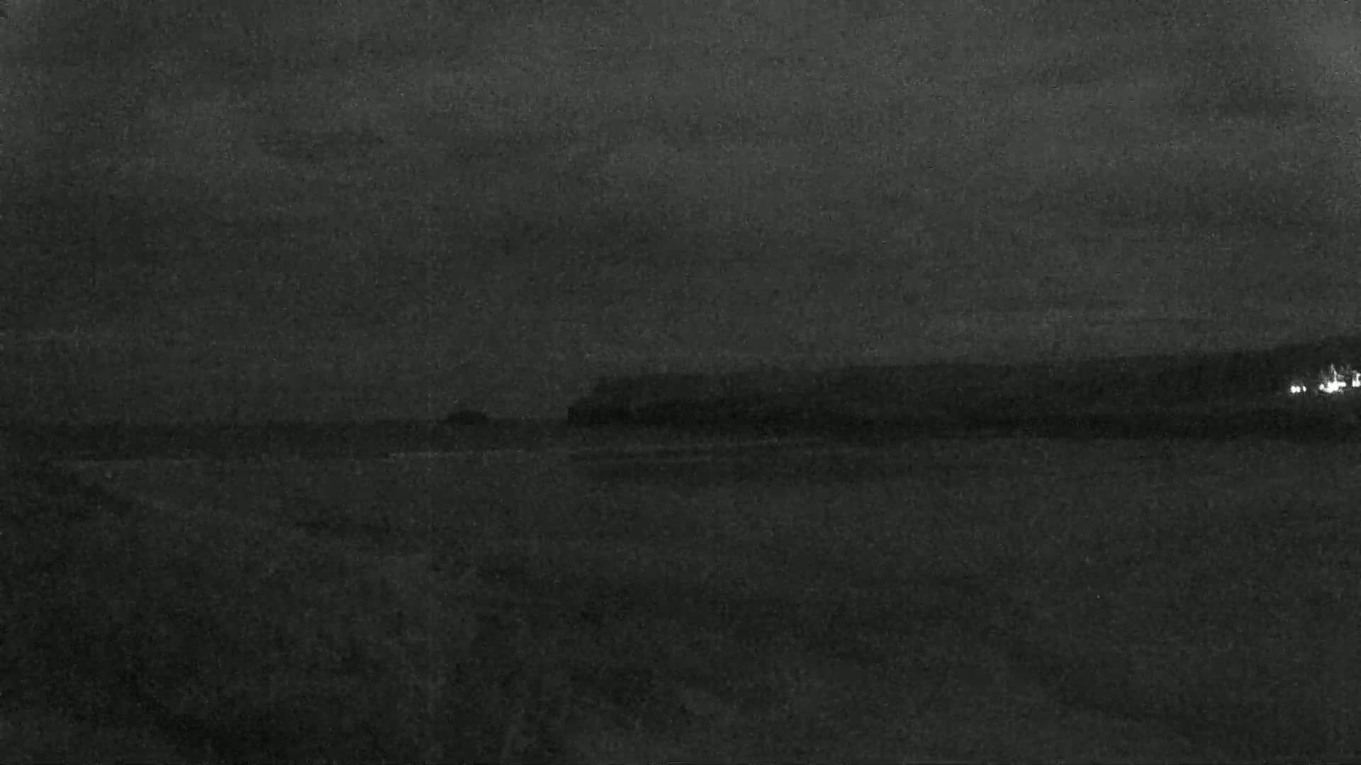 A dark, nighttime coastal scene shows a flat body of water meeting a dark shoreline under a cloudy sky.