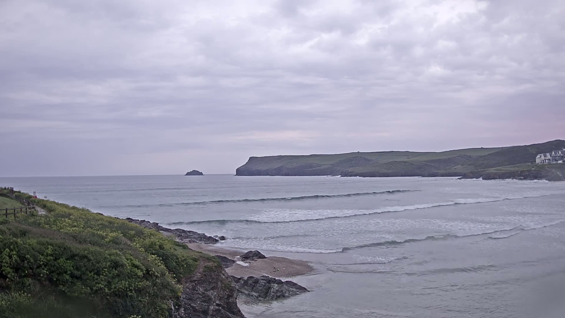 Gentle waves roll onto a sandy beach, bordered by green cliffs and a distant, rocky island under a cloudy sky.