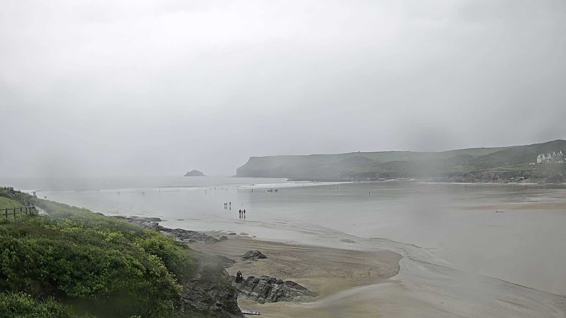 A wide, sandy beach stretches out to a calm sea under a foggy, overcast sky, with a few people walking along the shore.