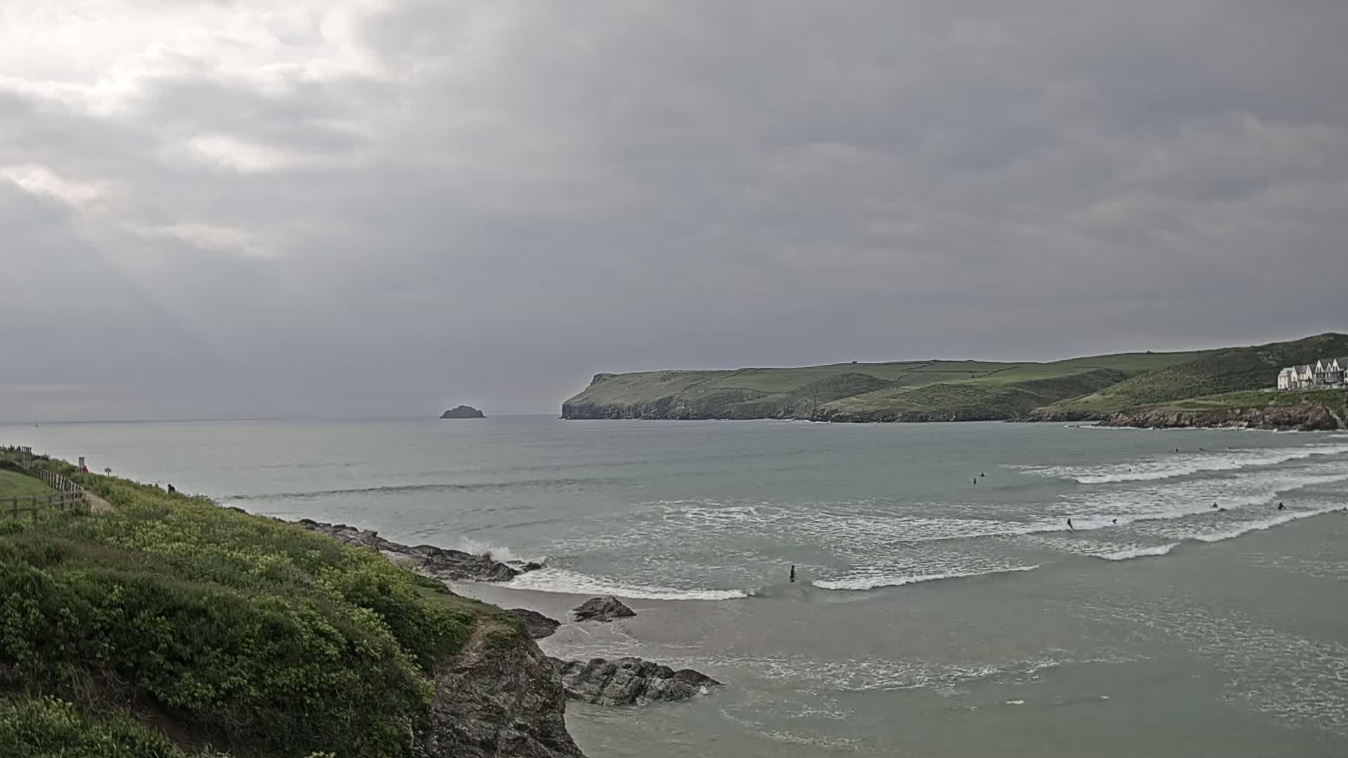 A coastal scene under an overcast sky shows a calm bay with several surfers, green hills, and a small island in the distance.
