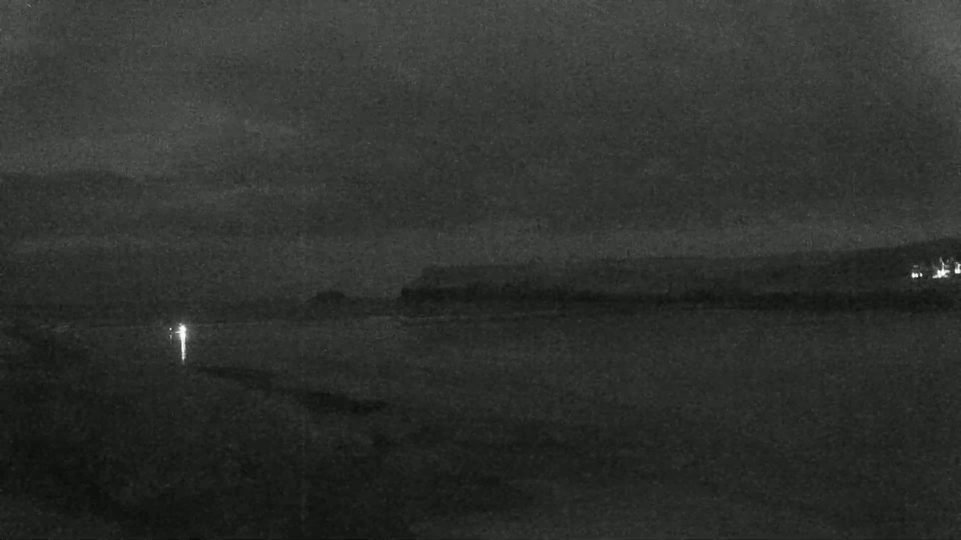 A dark, nighttime coastal scene shows a body of water in the foreground and a distant shoreline under a cloudy sky.