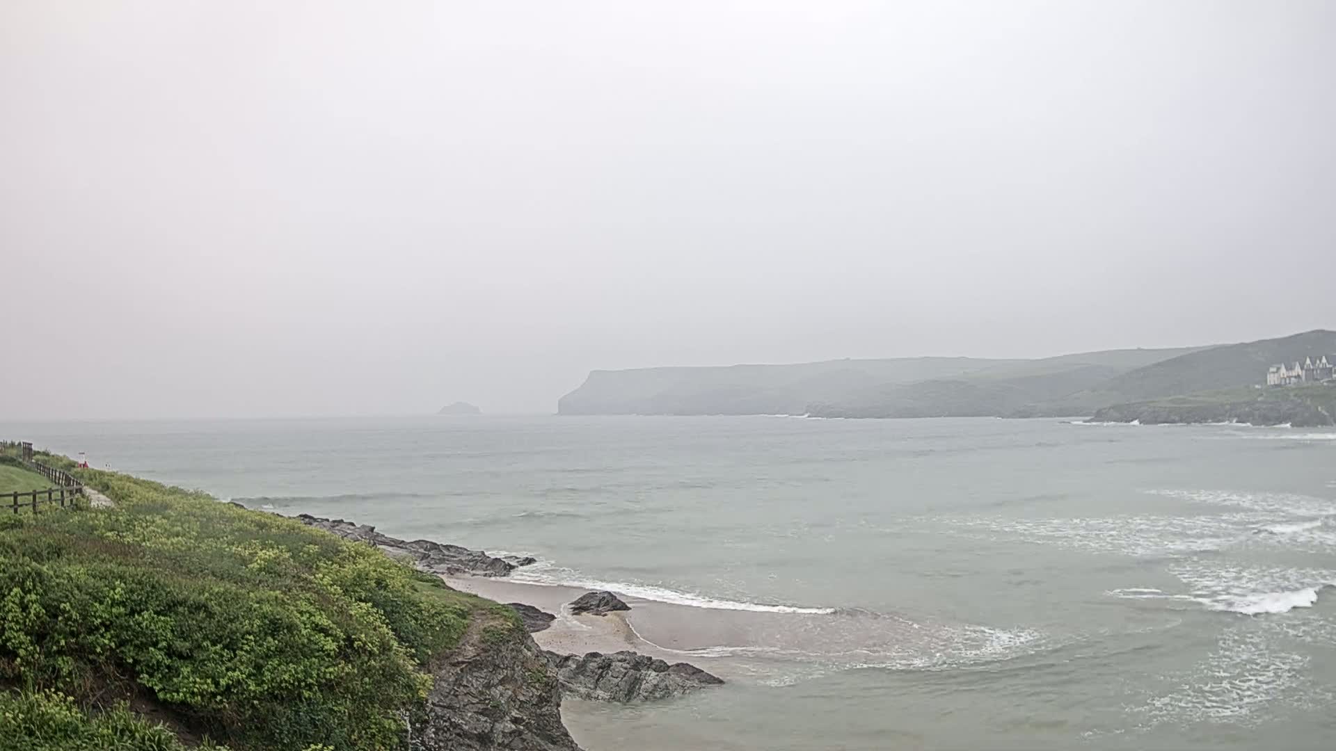 A hazy, overcast day reveals a coastal scene with a sandy beach nestled between rocky cliffs, overlooking a calm sea with gentle waves, and distant headlands under a muted sky.