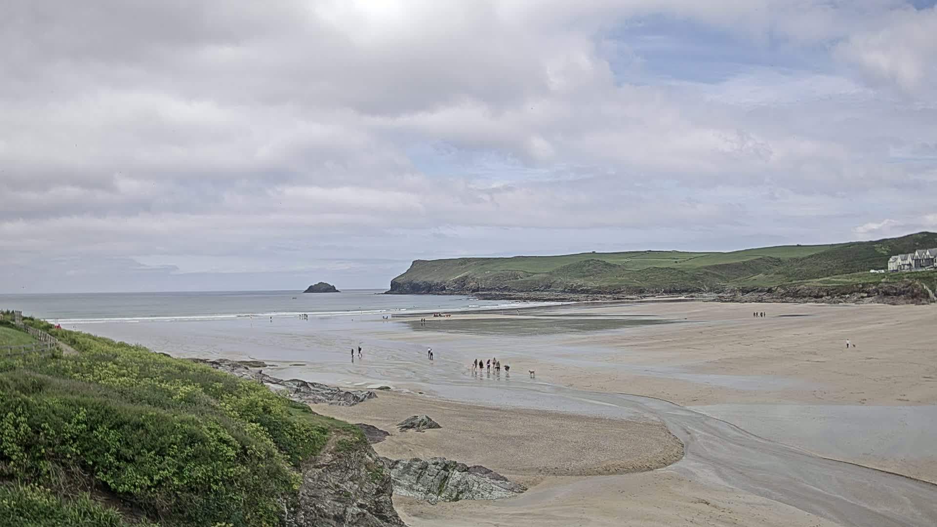 A wide sandy beach stretches out under a partly cloudy sky, with people walking on the sand and a small island visible in the distance.