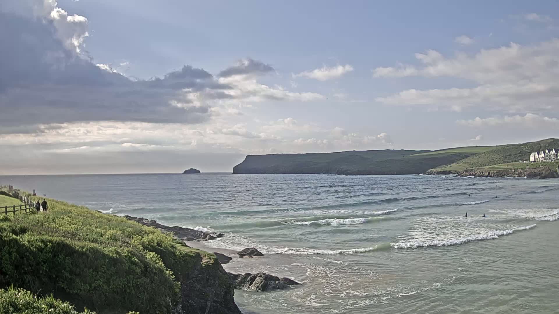 Two people walk along a grassy coastal path overlooking a bay with gentle waves under a partly cloudy sky.