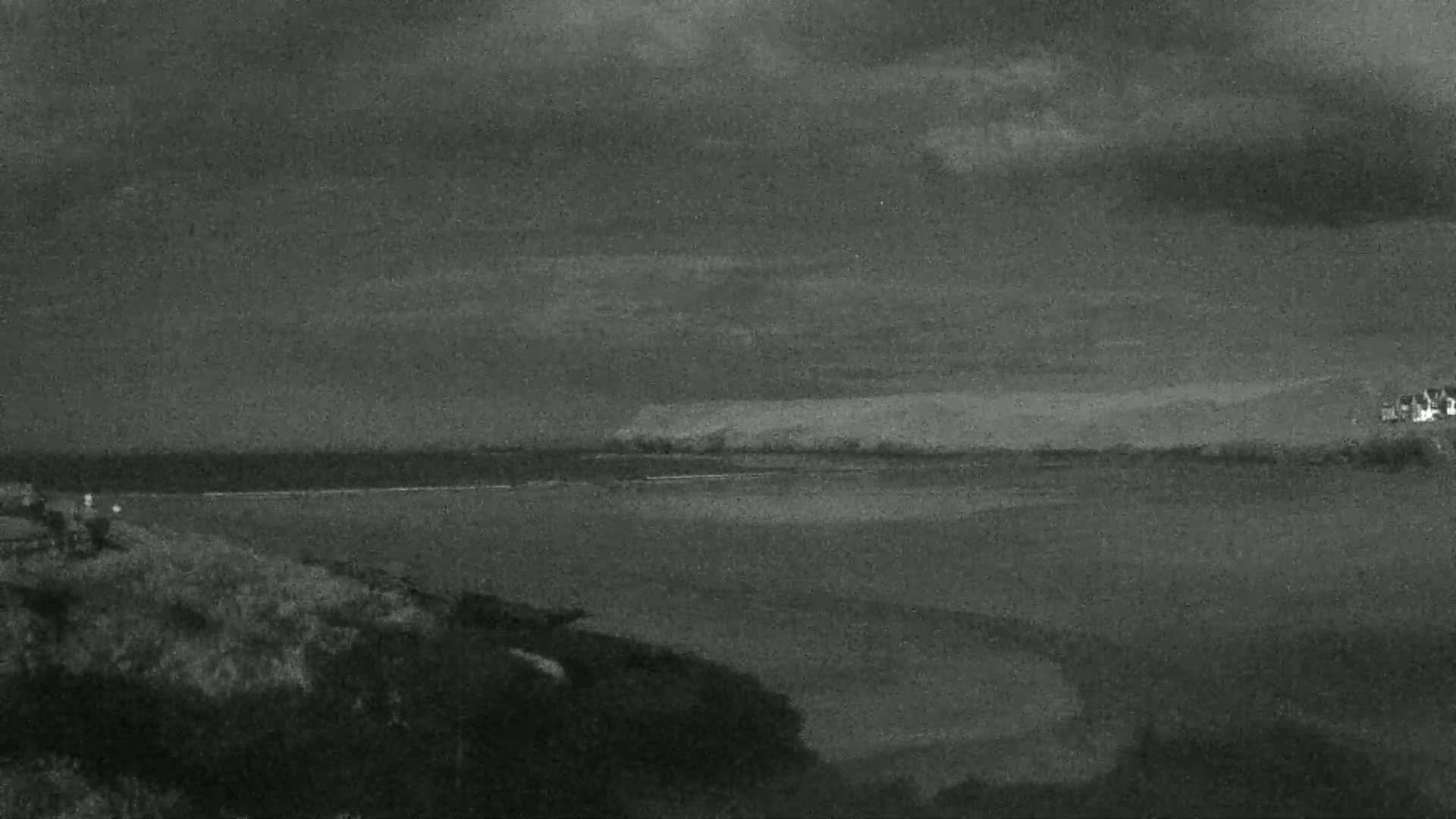 A dark, overcast sky hangs over a calm body of water, with land visible in the distance and vegetation in the foreground.