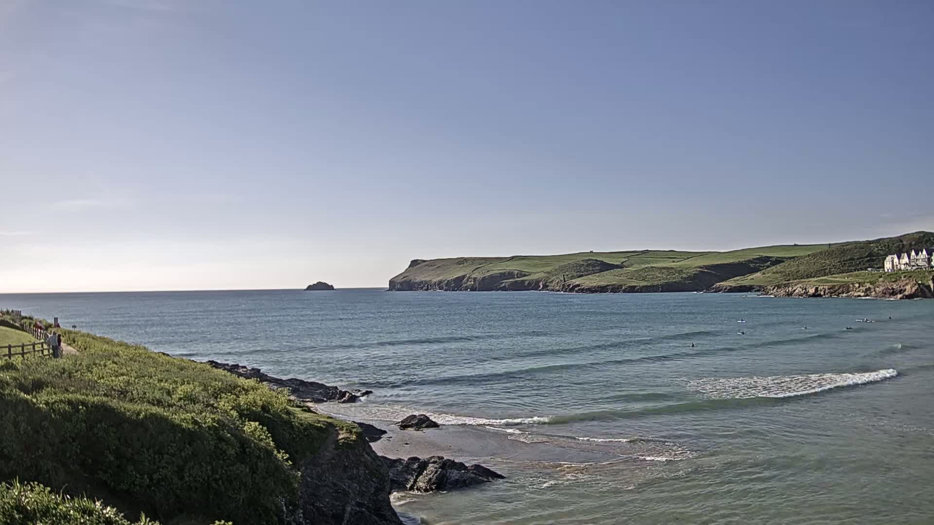 A clear, sunny day reveals a coastal scene with gentle waves lapping a sandy beach, a rocky cove, and a green, hilly landscape in the distance.