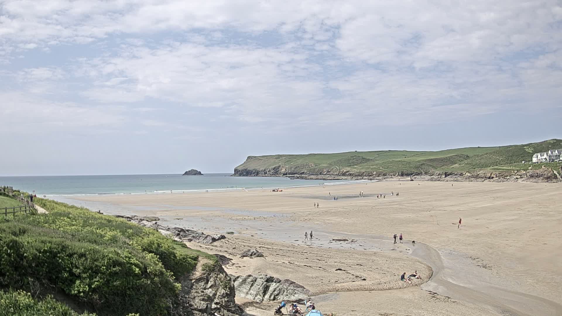 A sandy beach stretches out to a calm ocean under a partly cloudy sky, with a few people scattered across the sand and a small stream cutting through it.