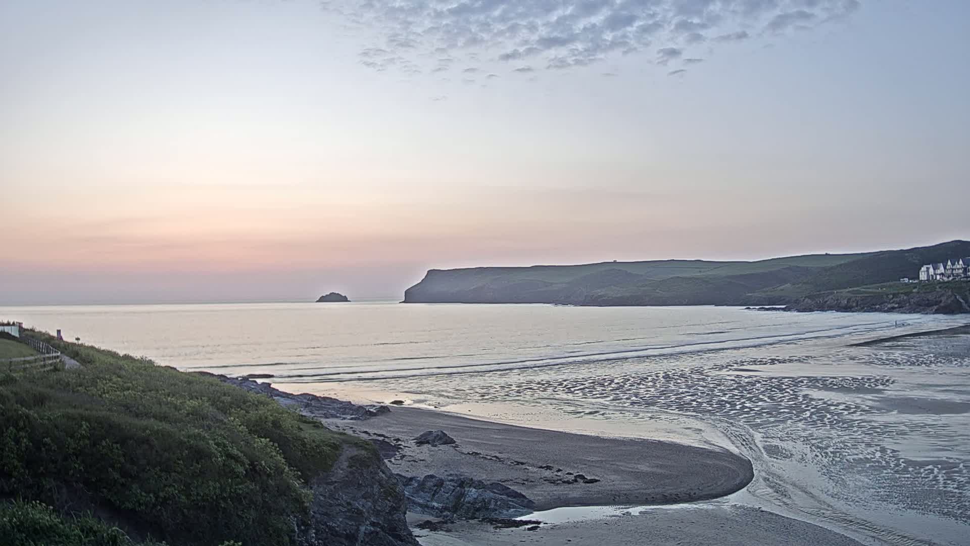 A tranquil beach scene at sunset shows a calm ocean with a small island and distant headland under a pale sky.