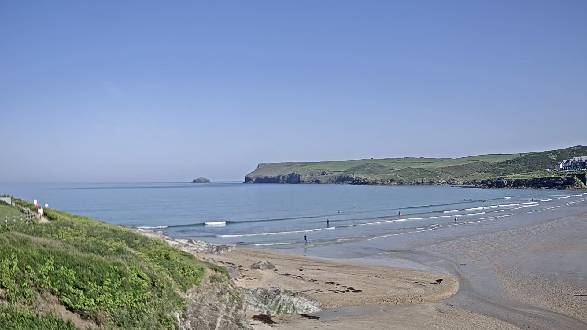 A sandy beach with gentle waves stretches out to a calm ocean under a clear blue sky, with a few people enjoying the water and a distant headland in view.
