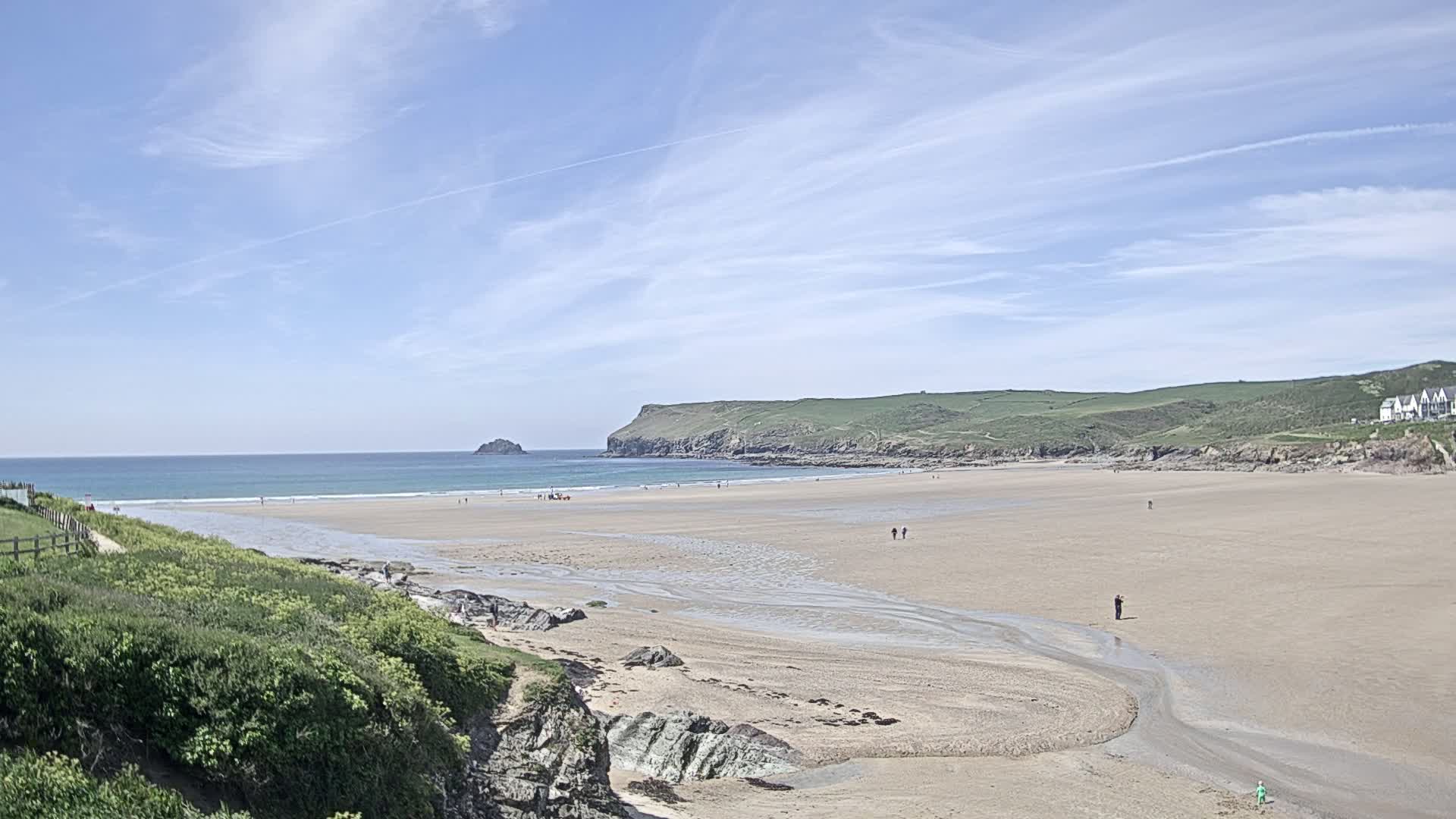 A sandy beach stretches out to a calm ocean under a mostly sunny sky, with green hills and a small island in the distance.