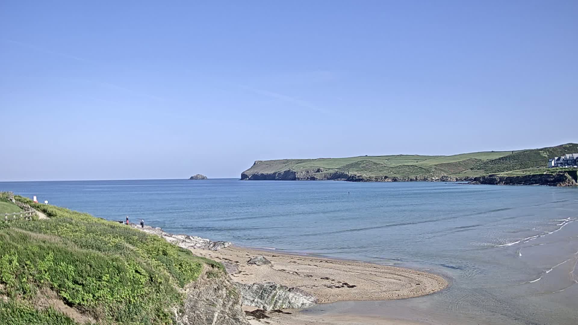 A sandy cove with calm, blue water under a clear blue sky is nestled between green hills and a rocky headland, with a few people visible on a path along the shore.