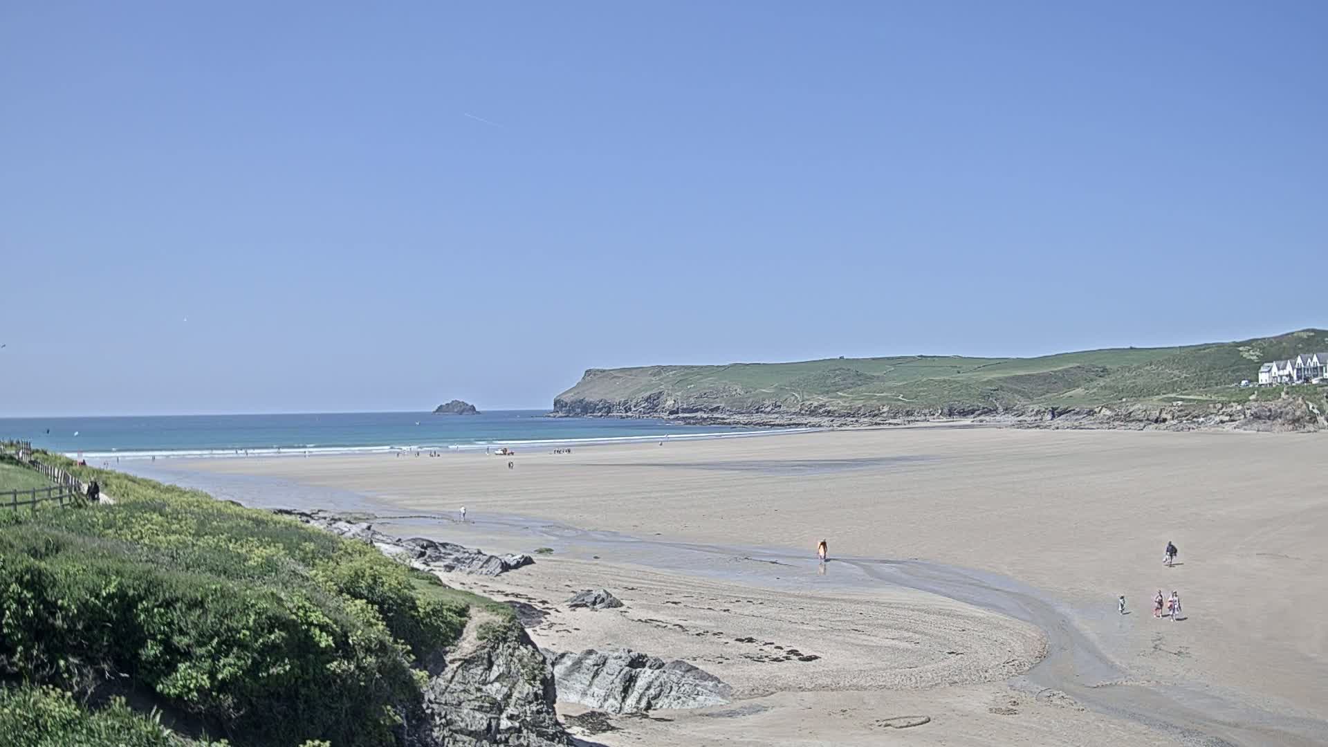 A wide sandy beach stretches out to a calm ocean under a clear blue sky, with a few people scattered across the sand and a small island visible in the distance.