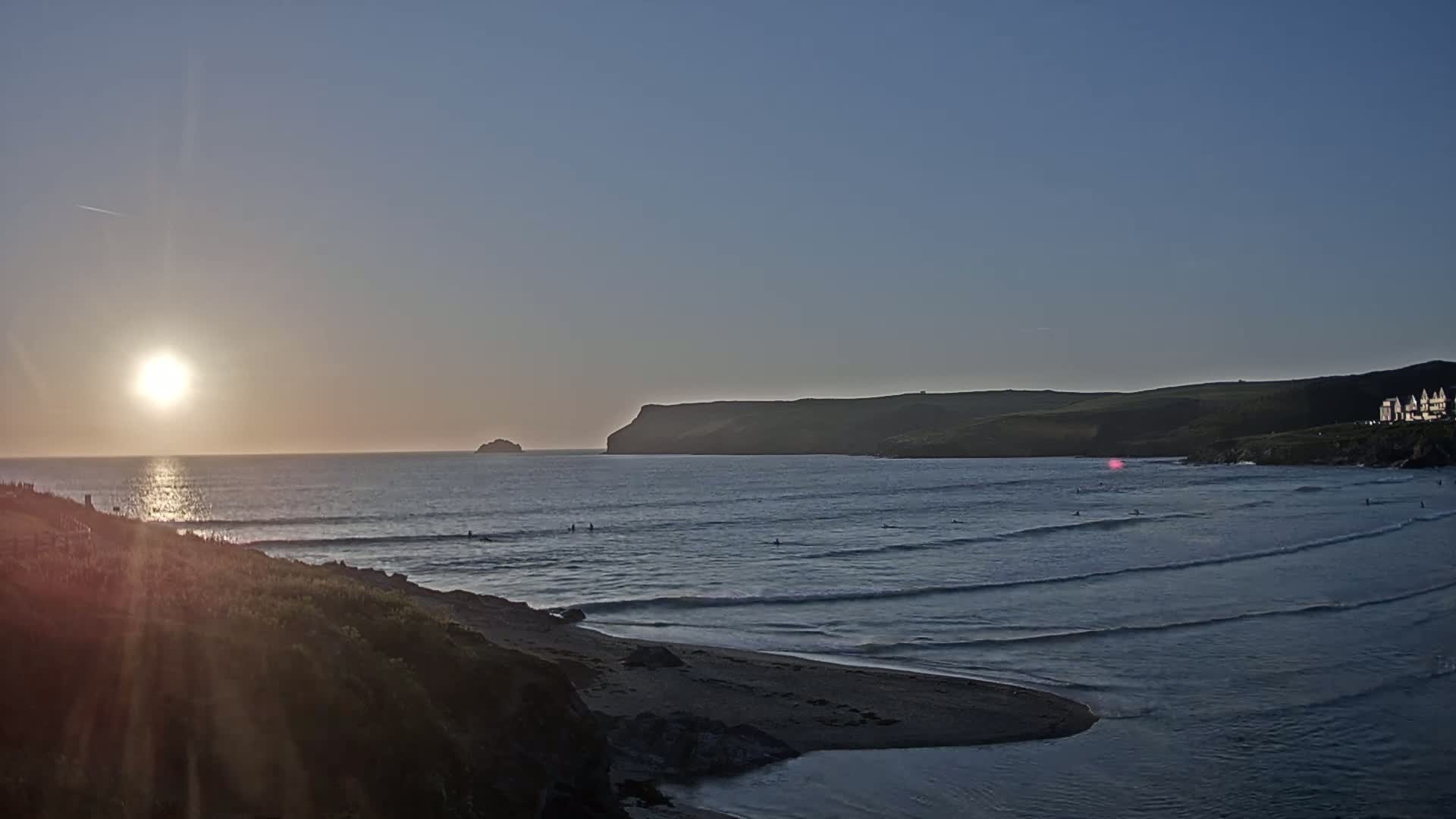A setting sun casts a warm glow over a calm ocean bay, where several people are surfing near a sandy shore and a rocky cliff.