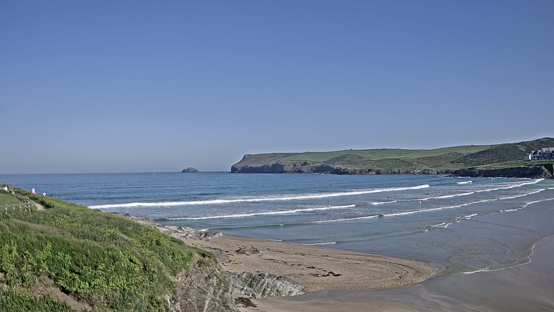 A sandy beach with gentle waves under a clear blue sky, featuring a rocky coastline and a distant headland.