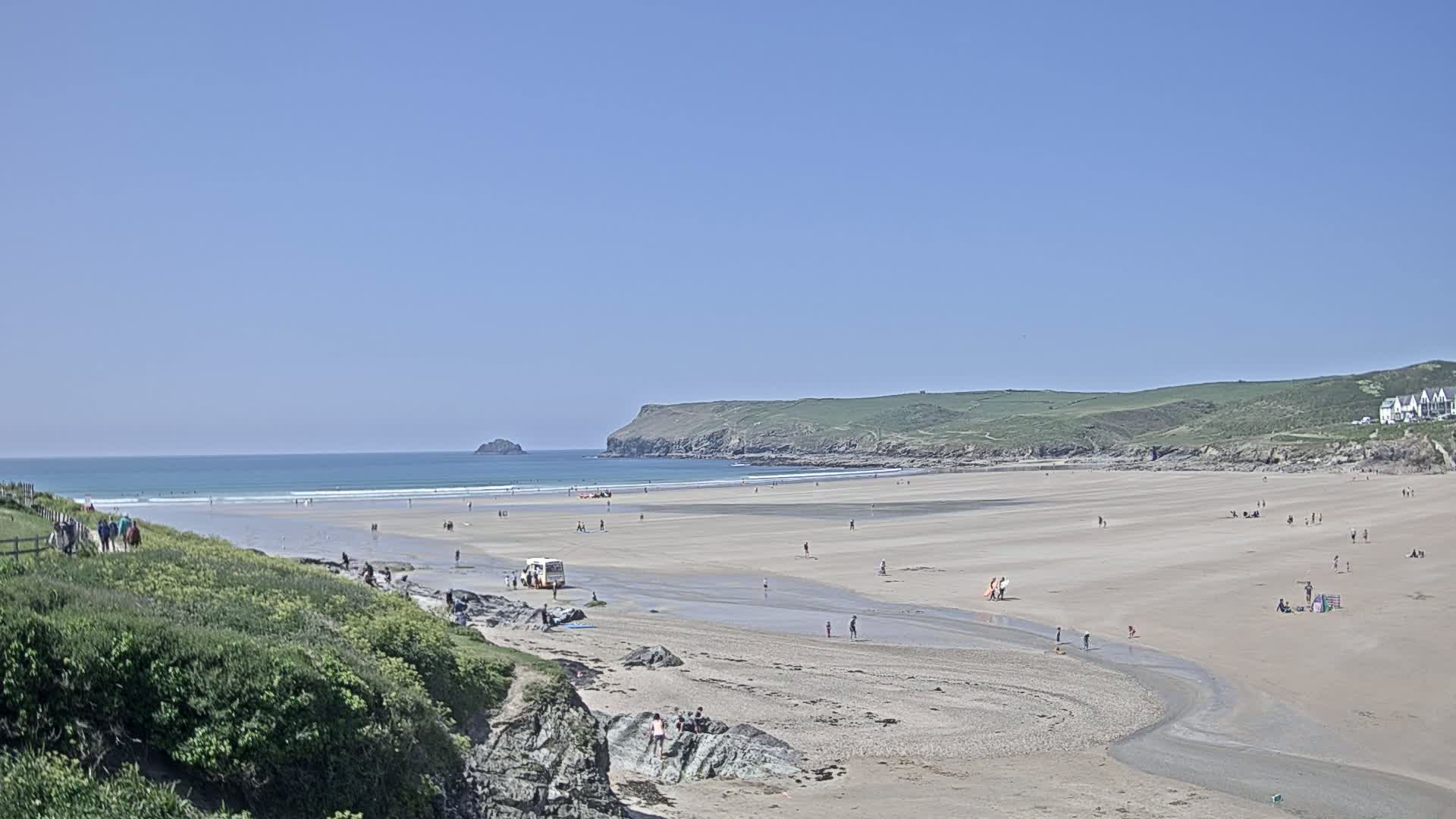 A sandy beach stretches out to a calm ocean under a clear blue sky, with a few people scattered across the sand and some surfers in the water.
