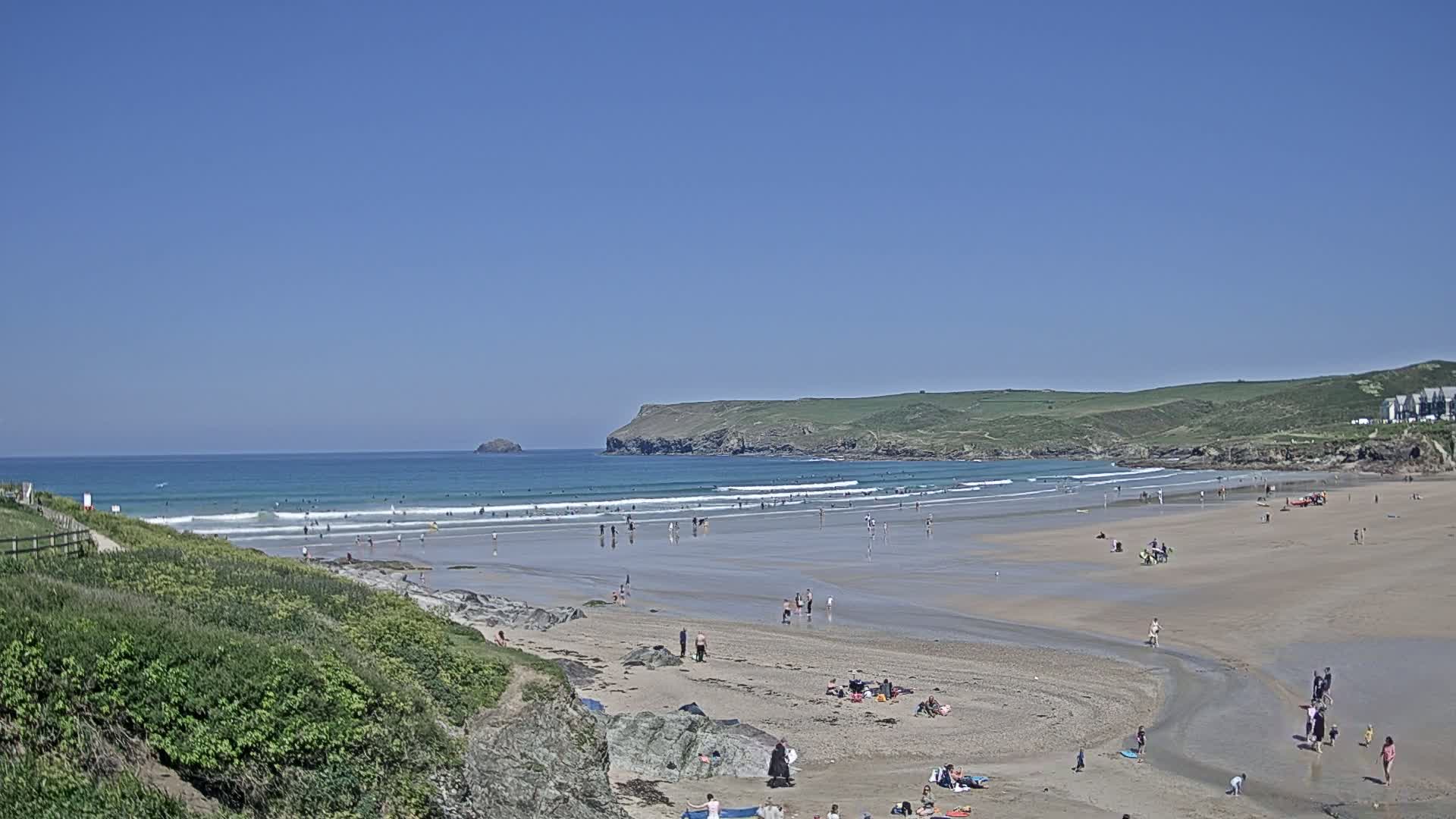 A sandy beach with many people on it and in the ocean under a clear blue sky is seen from a high vantage point.