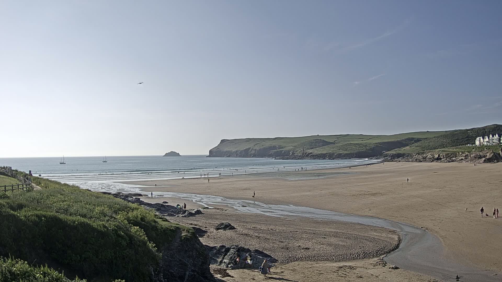 A sandy beach stretches out to a calm ocean under a clear, bright sky, with a few people scattered along the shore and a small stream flowing from the land to the sea.