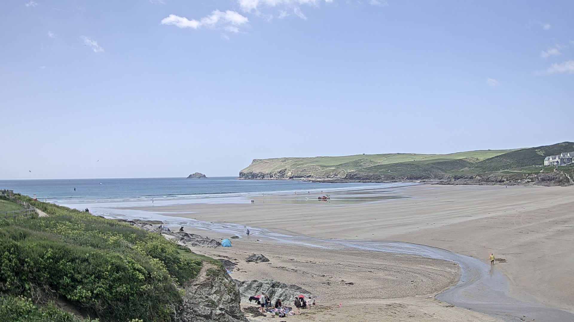 A sandy beach stretches out to a calm ocean under a mostly sunny sky, with a few people scattered on the sand and in the water, and green hills rising in the background.