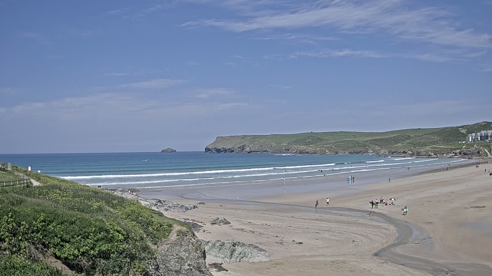 A sandy beach with rolling waves under a clear blue sky, showing several people enjoying the sunny weather.