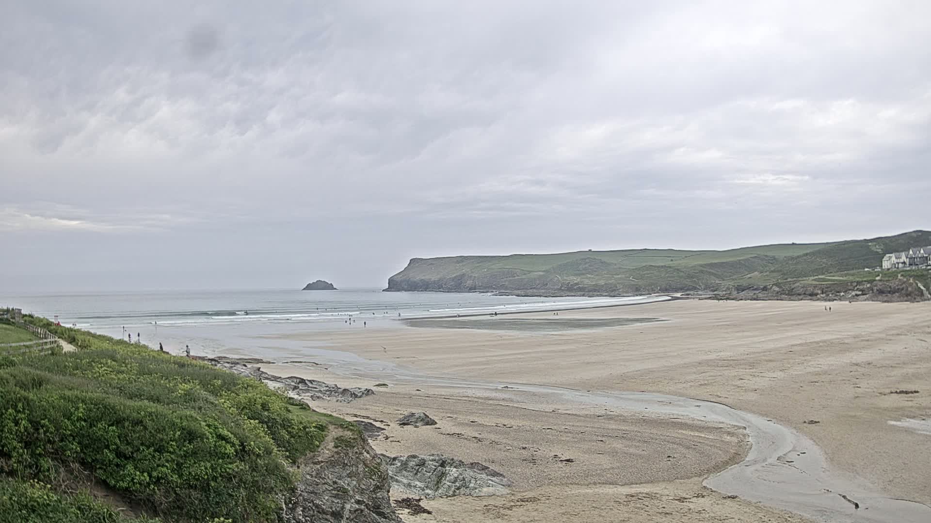 A wide sandy beach stretches out to a calm ocean under a cloudy sky, with a few people scattered on the sand and in the water.
