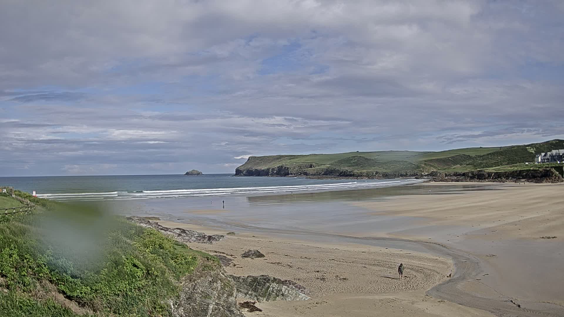 A sandy beach stretches out to a calm ocean under a partly cloudy sky, with small waves rolling in and a few people walking along the shore.