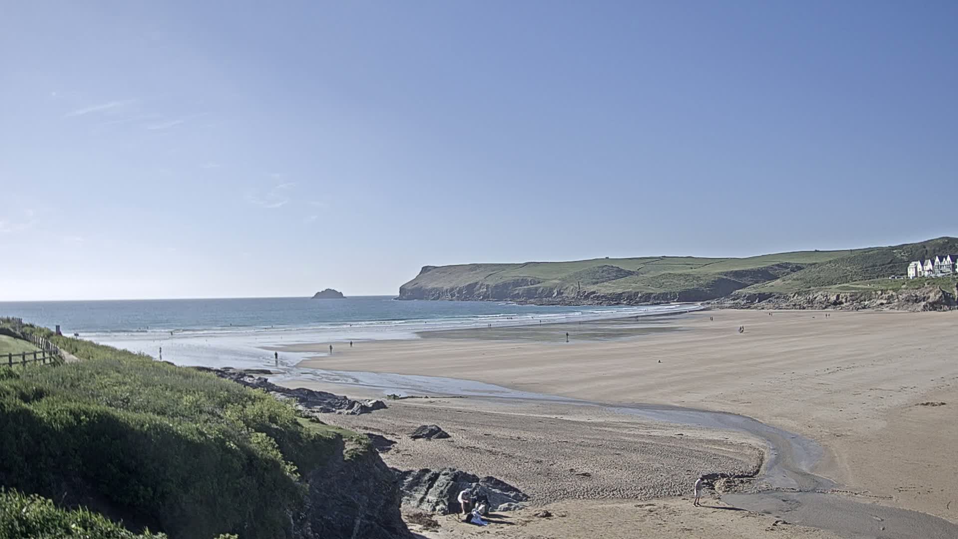 A sandy beach stretches out to a calm ocean under a clear blue sky, with a few people scattered on the shore and a small island visible in the distance.