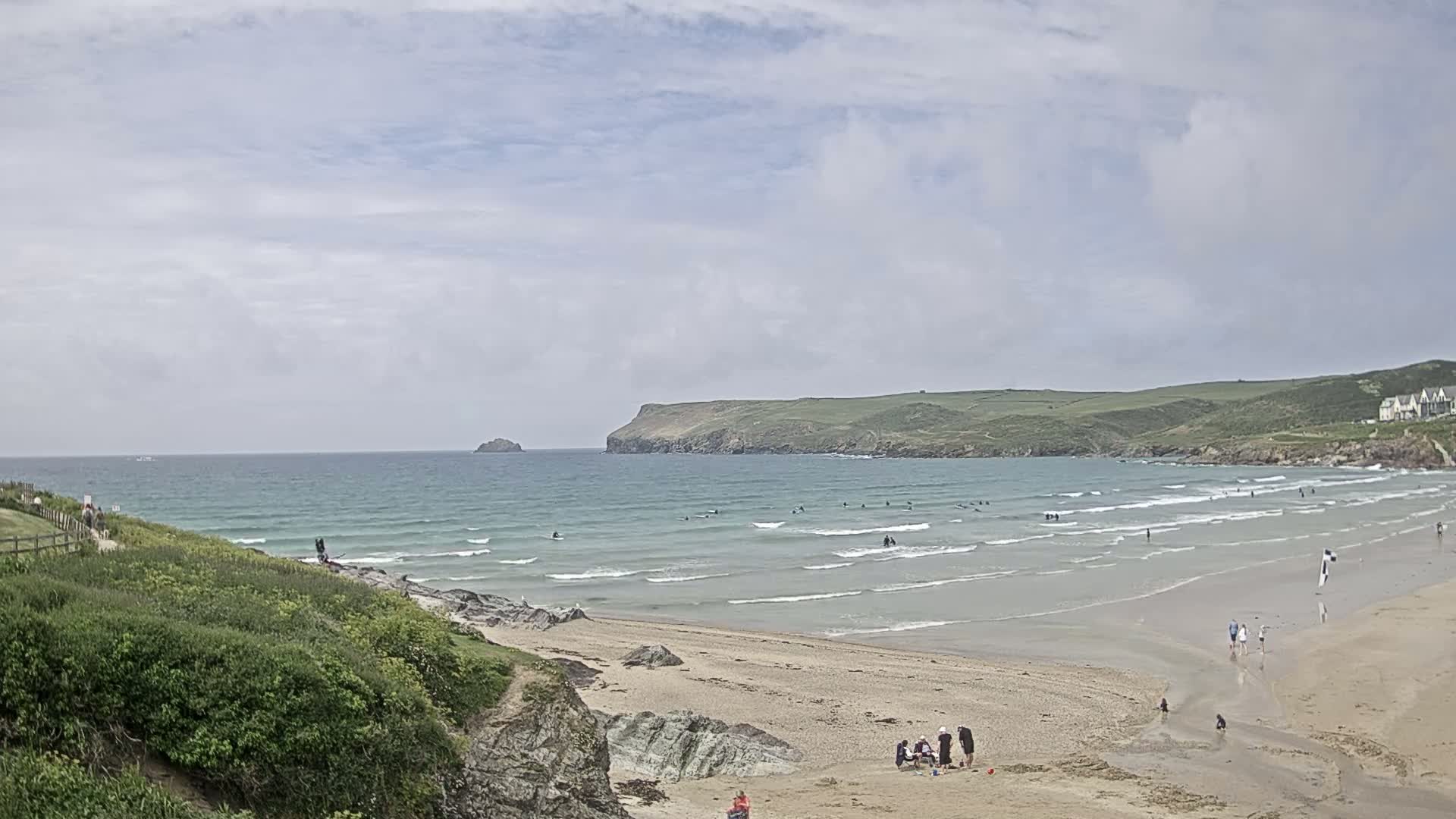 A sandy beach with several people on it, under a mostly cloudy sky, has gentle waves breaking in the ocean and surfers in the water.