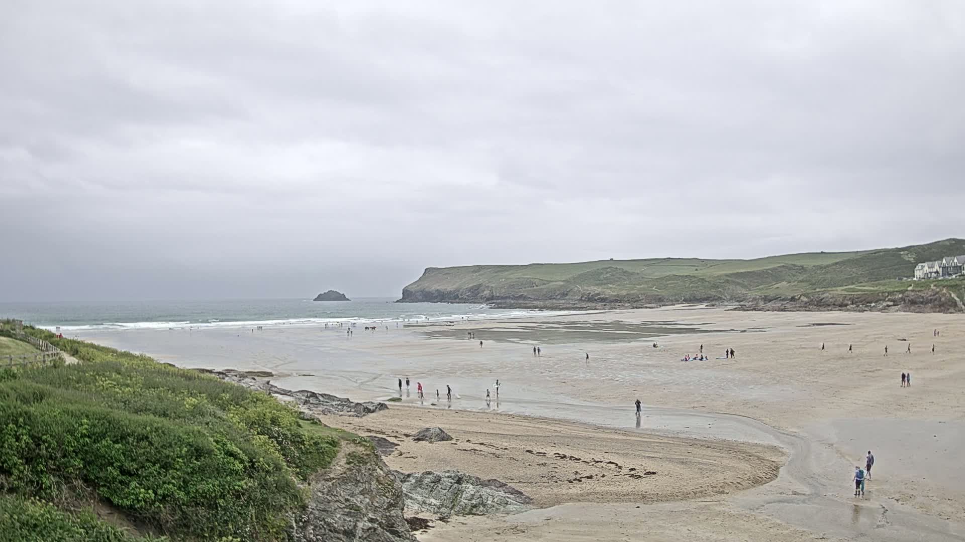 A wide sandy beach stretches out under an overcast sky, with scattered people enjoying the shore and a calm ocean in the background.
