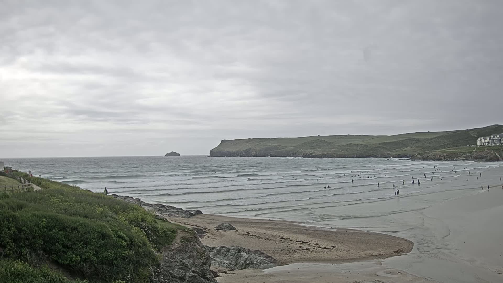 A sandy beach with gentle waves and many people in the water is seen under an overcast sky, with green hills and a small island in the distance.