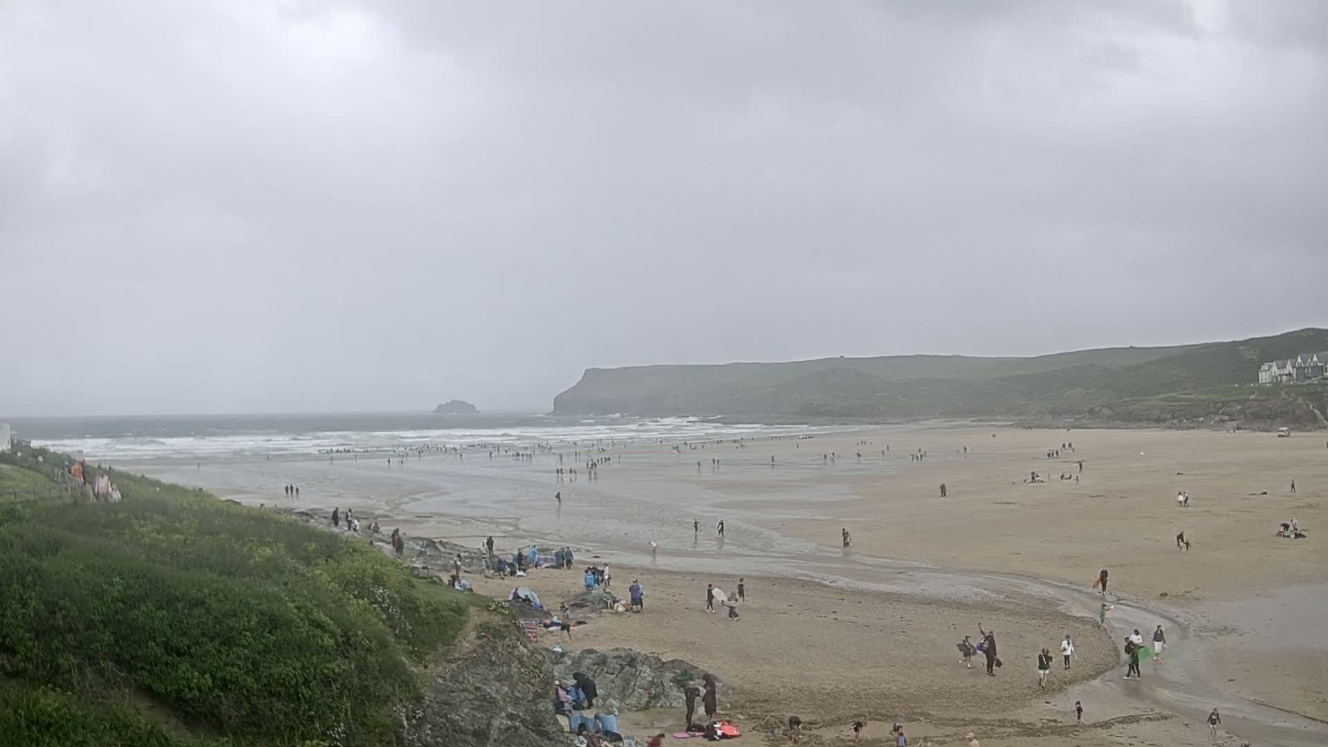 A wide sandy beach with many people scattered across it under an overcast sky, with waves breaking in the ocean beyond.