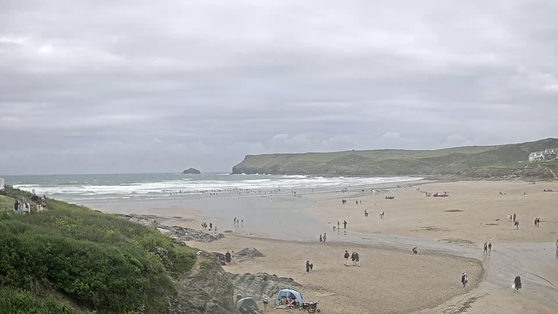 A wide sandy beach with many people on it and small waves in the ocean under an overcast sky.