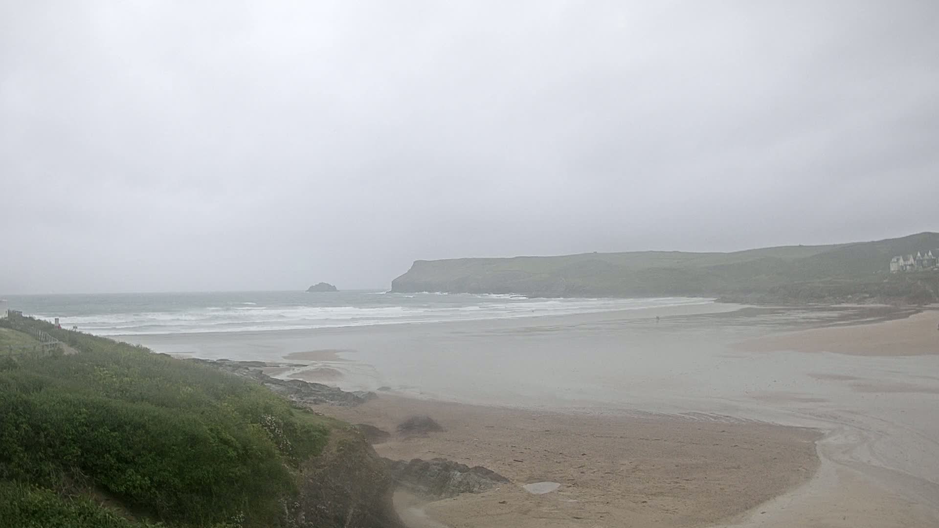 A hazy, overcast day reveals a sandy beach, a calm ocean with small waves, and a distant, verdant headland.