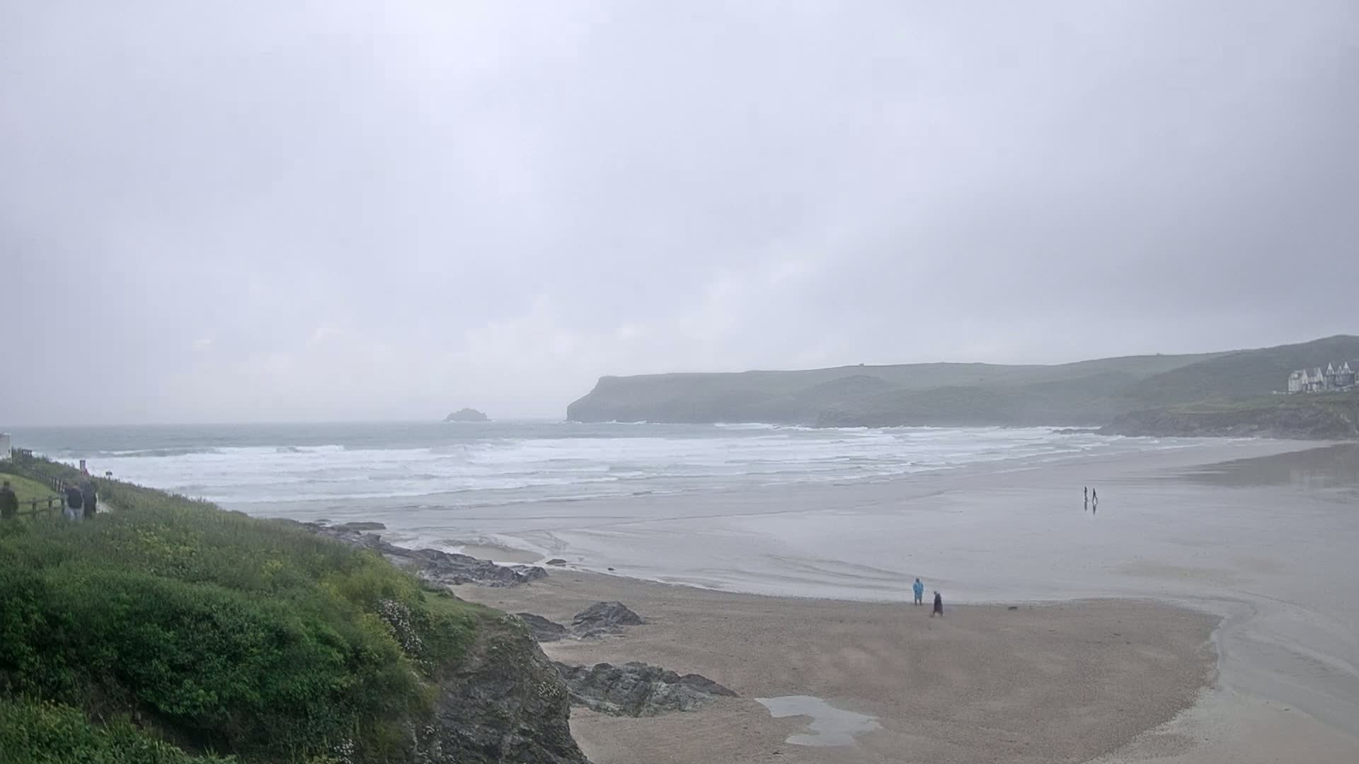 A wide sandy beach with small waves under an overcast sky, a few people are visible walking on the beach and along a cliffside path.
