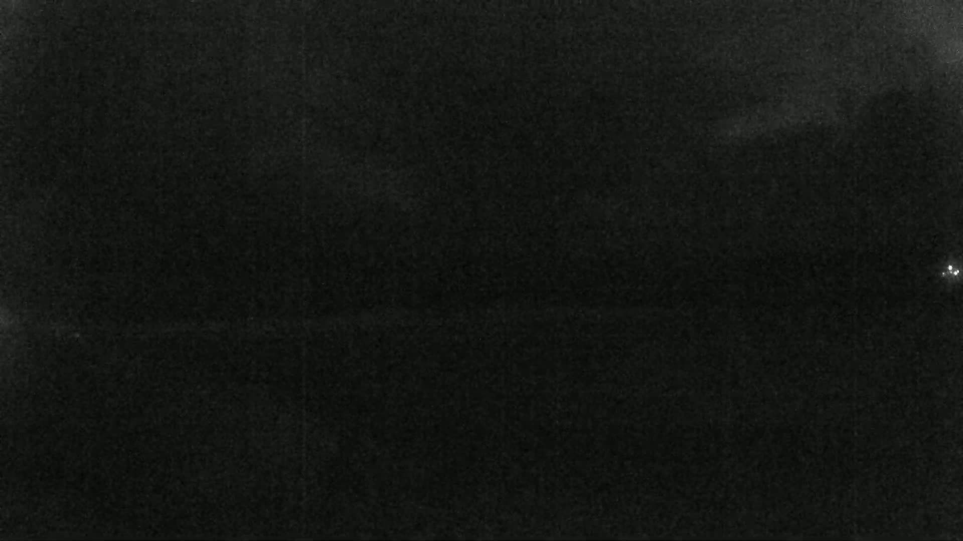 A dark, grainy image shows a mostly black scene with a few faint lights visible in the distance at night.