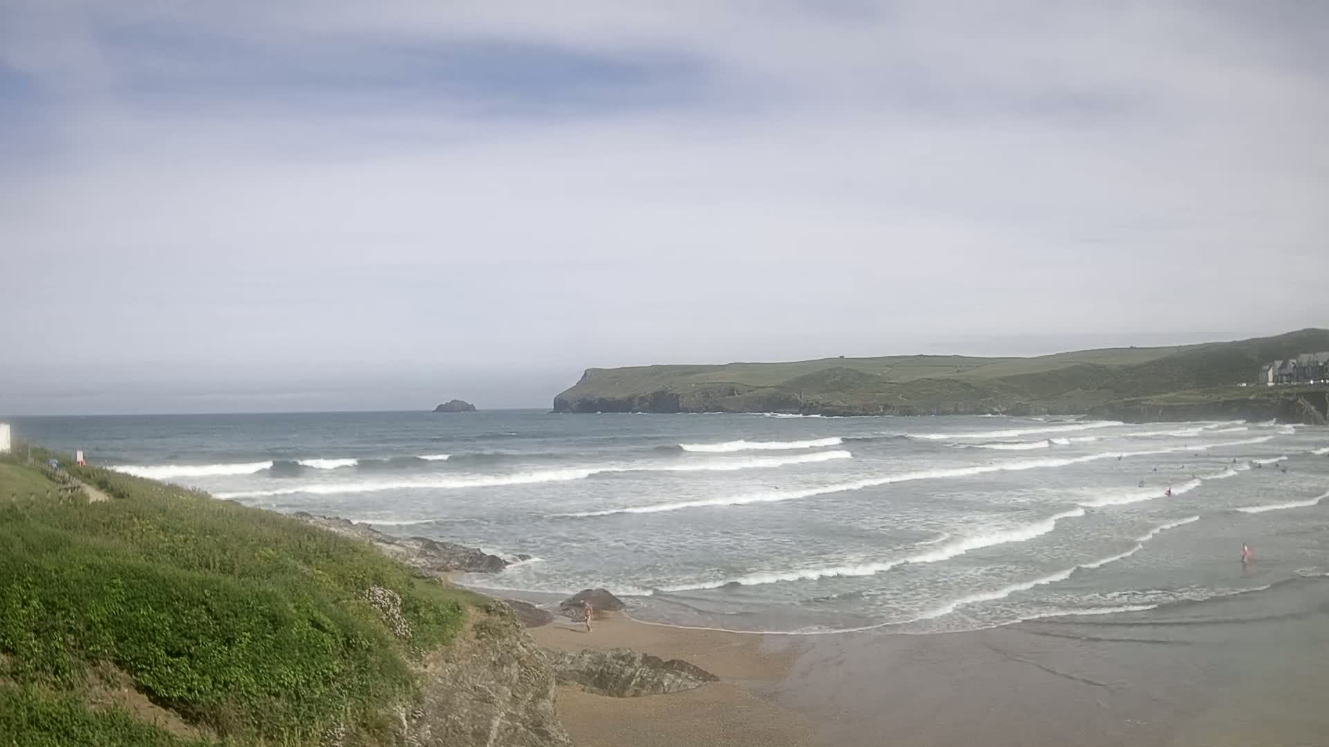 A sandy beach with rolling waves under a partly cloudy sky, featuring surfers in the water and green hills in the background.