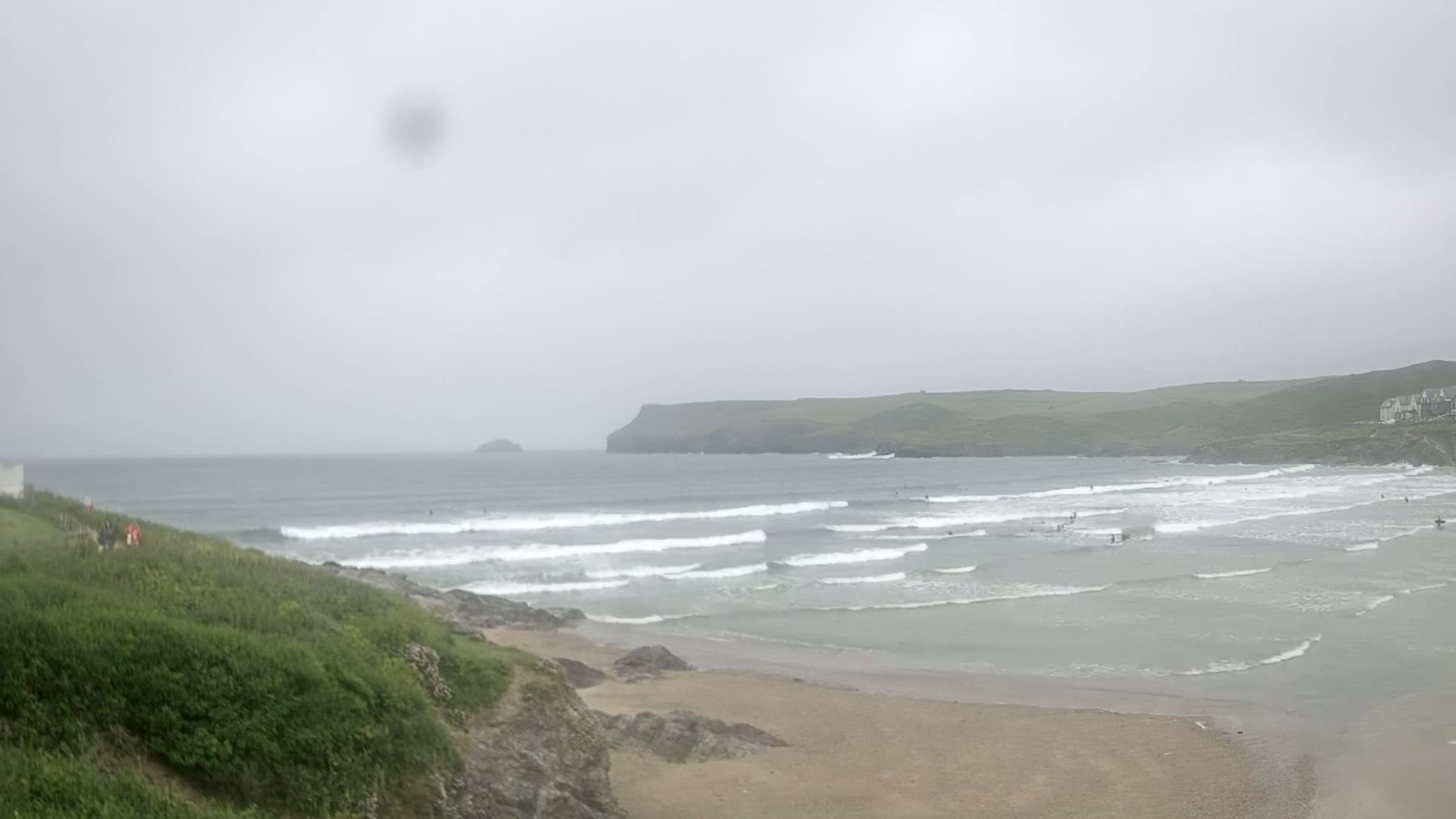 A sandy beach with small waves under an overcast sky, with a few surfers in the water and green hills in the background.