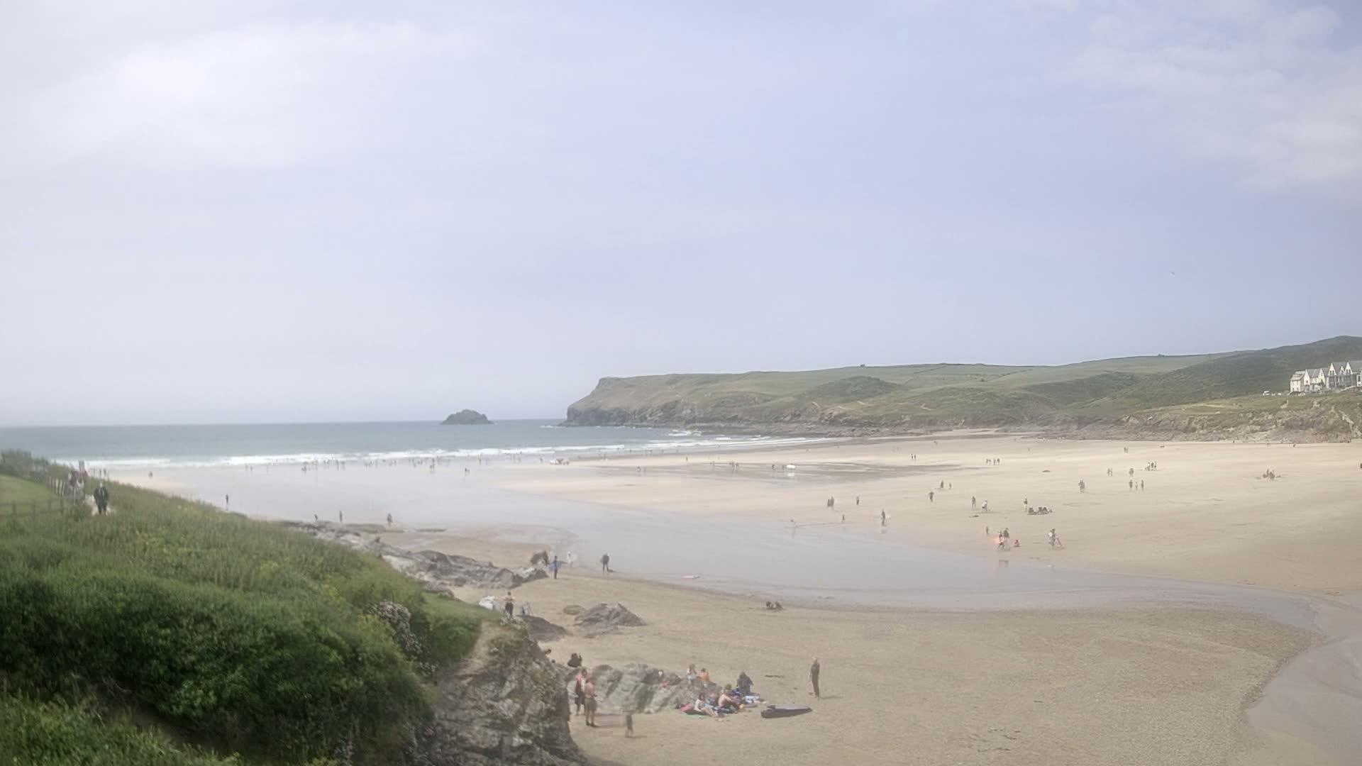 A wide sandy beach with a calm ocean, dotted with people, stretches under a cloudy sky.