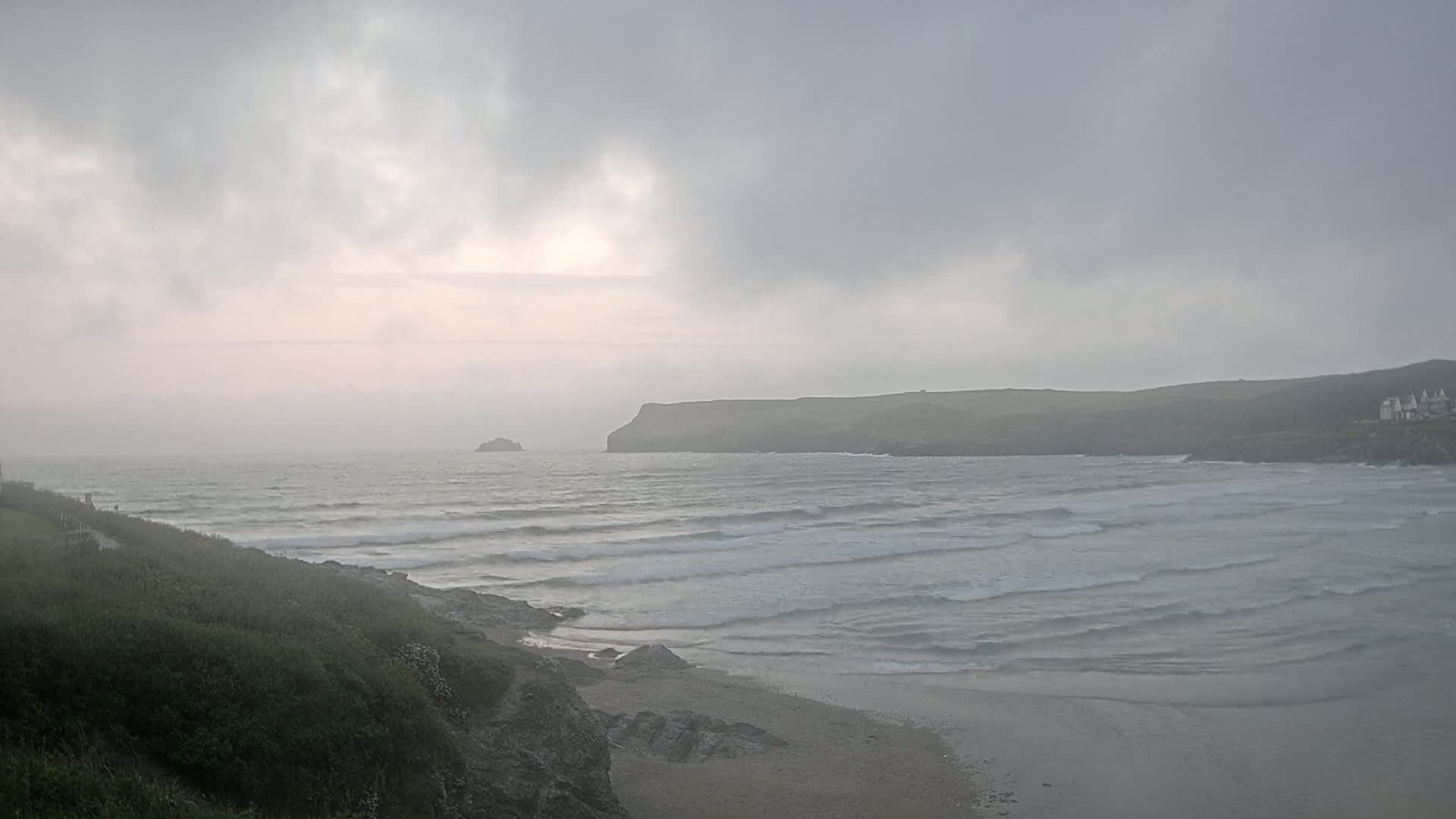 A hazy, overcast day reveals a coastal scene with rolling waves lapping a sandy beach, a small island in the distance, and a headland in the background.