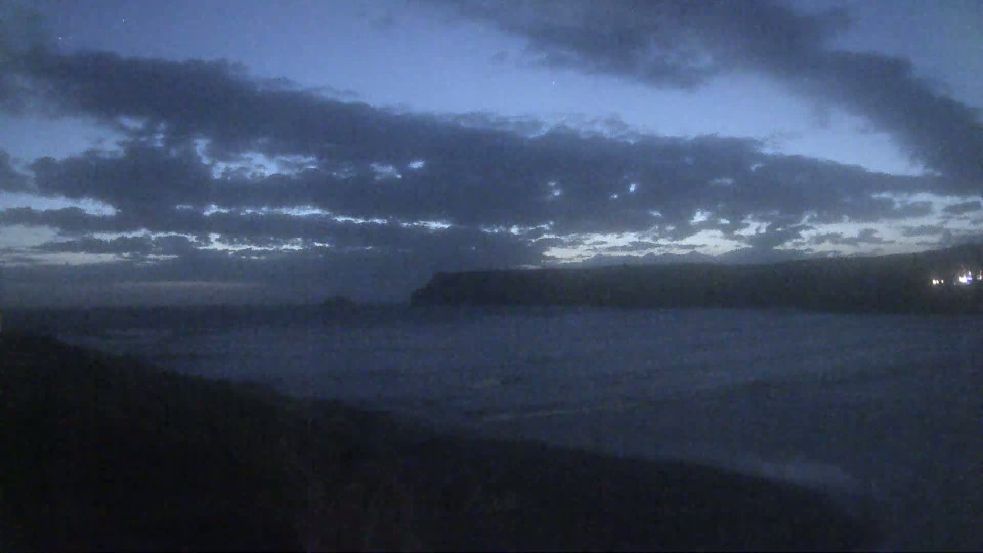 A dark, cloudy, pre-dawn sky hangs over a calm, misty ocean meeting a distant, silhouetted coastline.