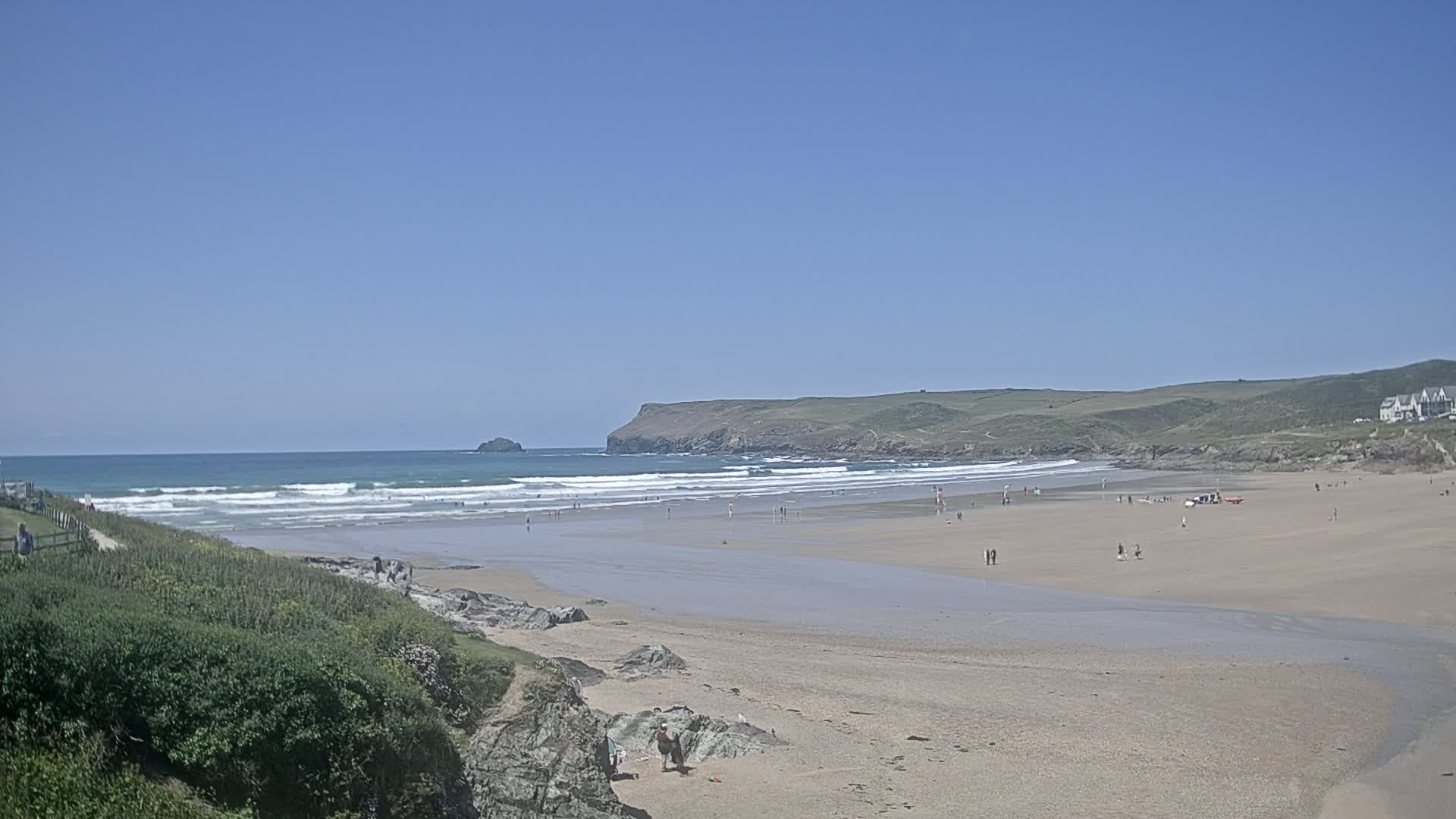 A wide sandy beach with gentle waves under a clear blue sky, sparsely populated with people.