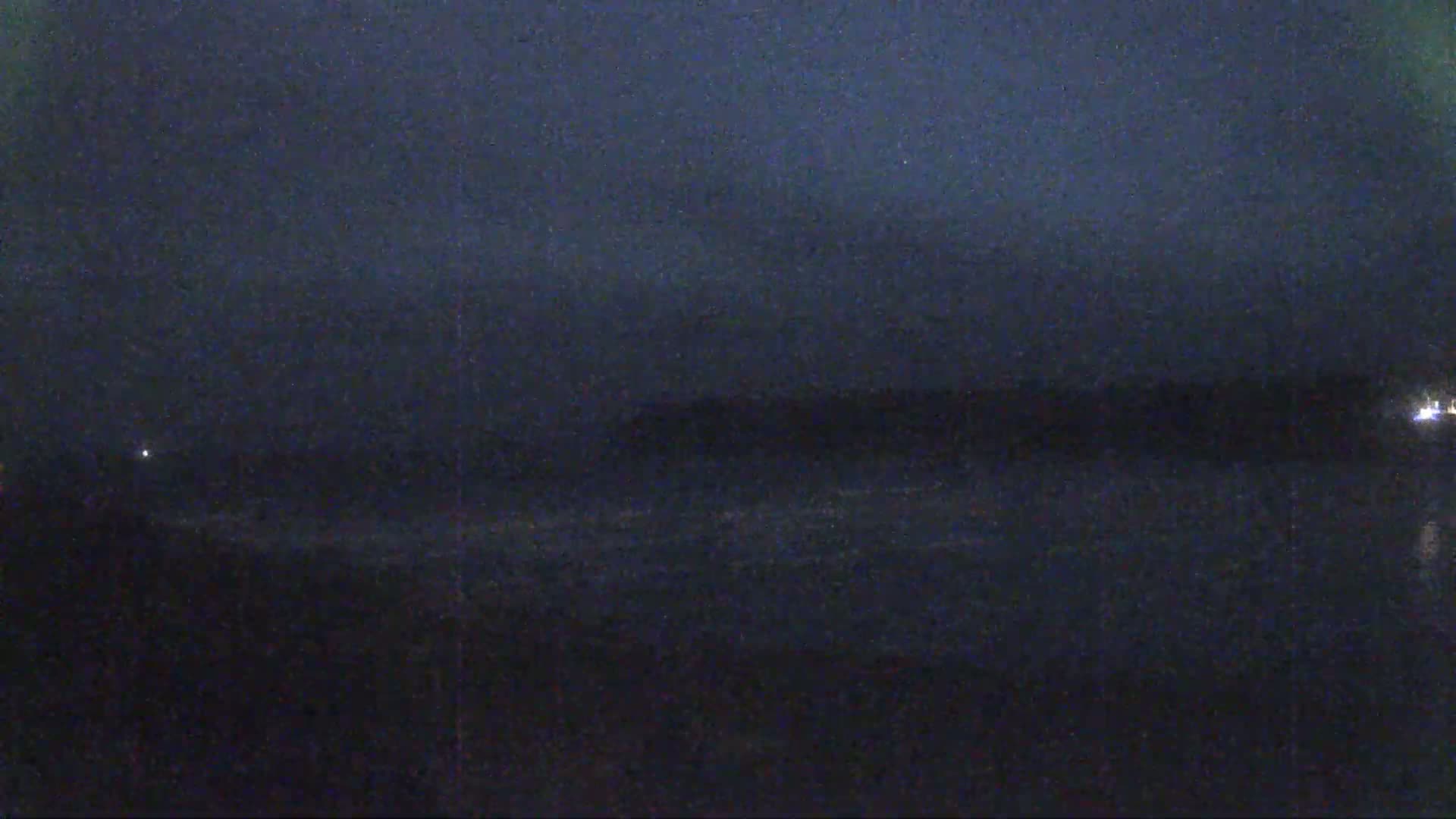 A dark, nighttime scene shows a body of water with a distant shoreline under a mostly clear, dark sky.