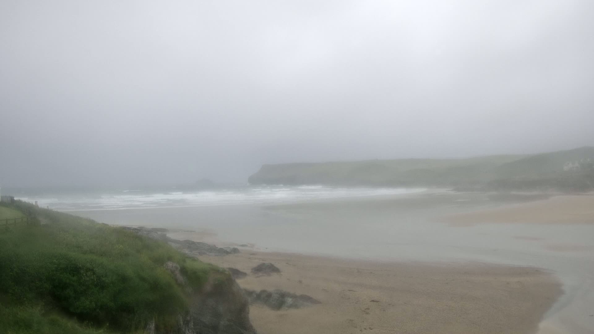 A foggy coastal scene shows a sandy beach meeting a calm bay, with a grassy headland visible in the distance and waves breaking gently on the shore.