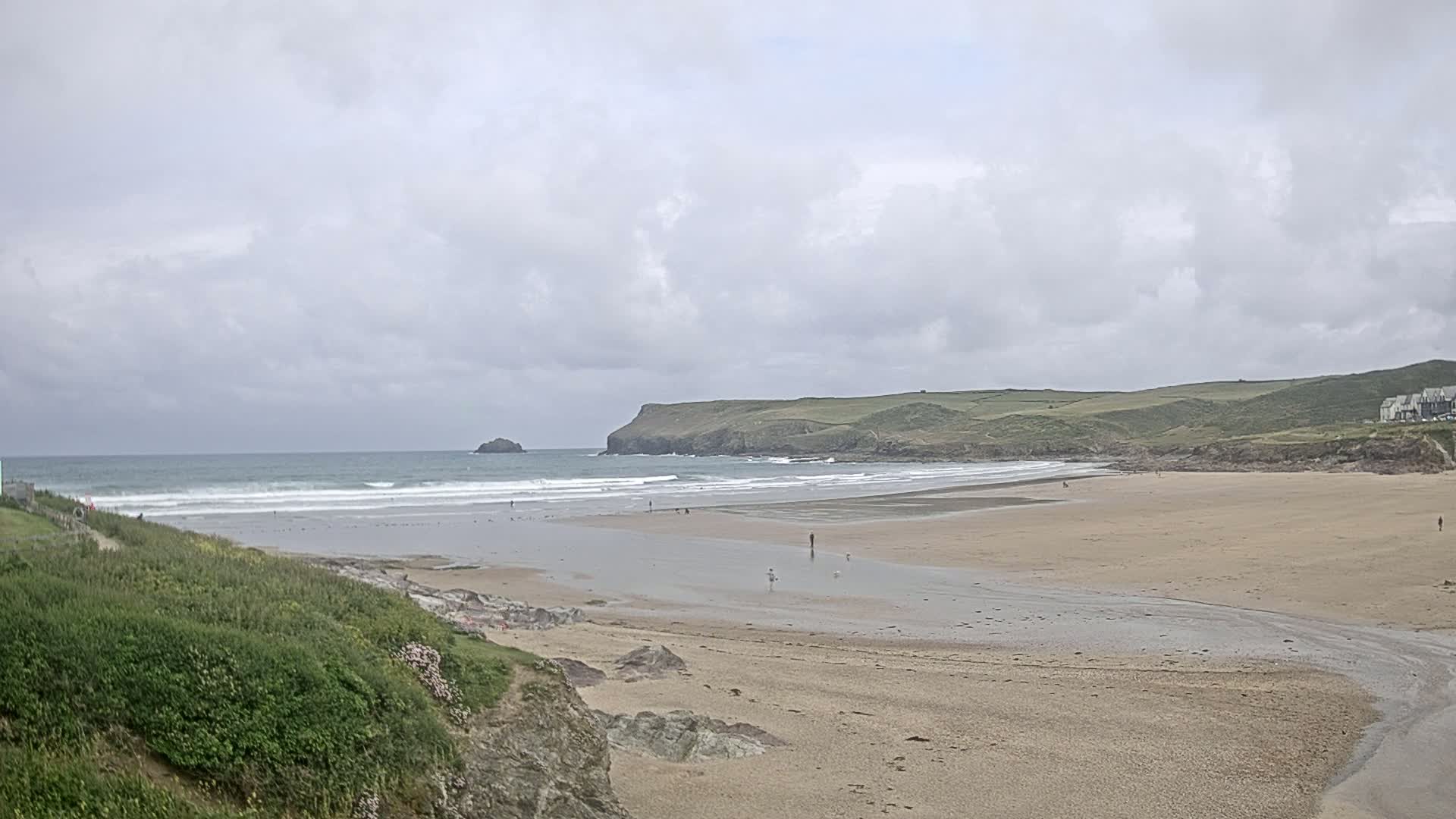 A sandy beach stretches out to a gently waving ocean under a cloudy sky, with a small island and grassy headland visible in the distance.