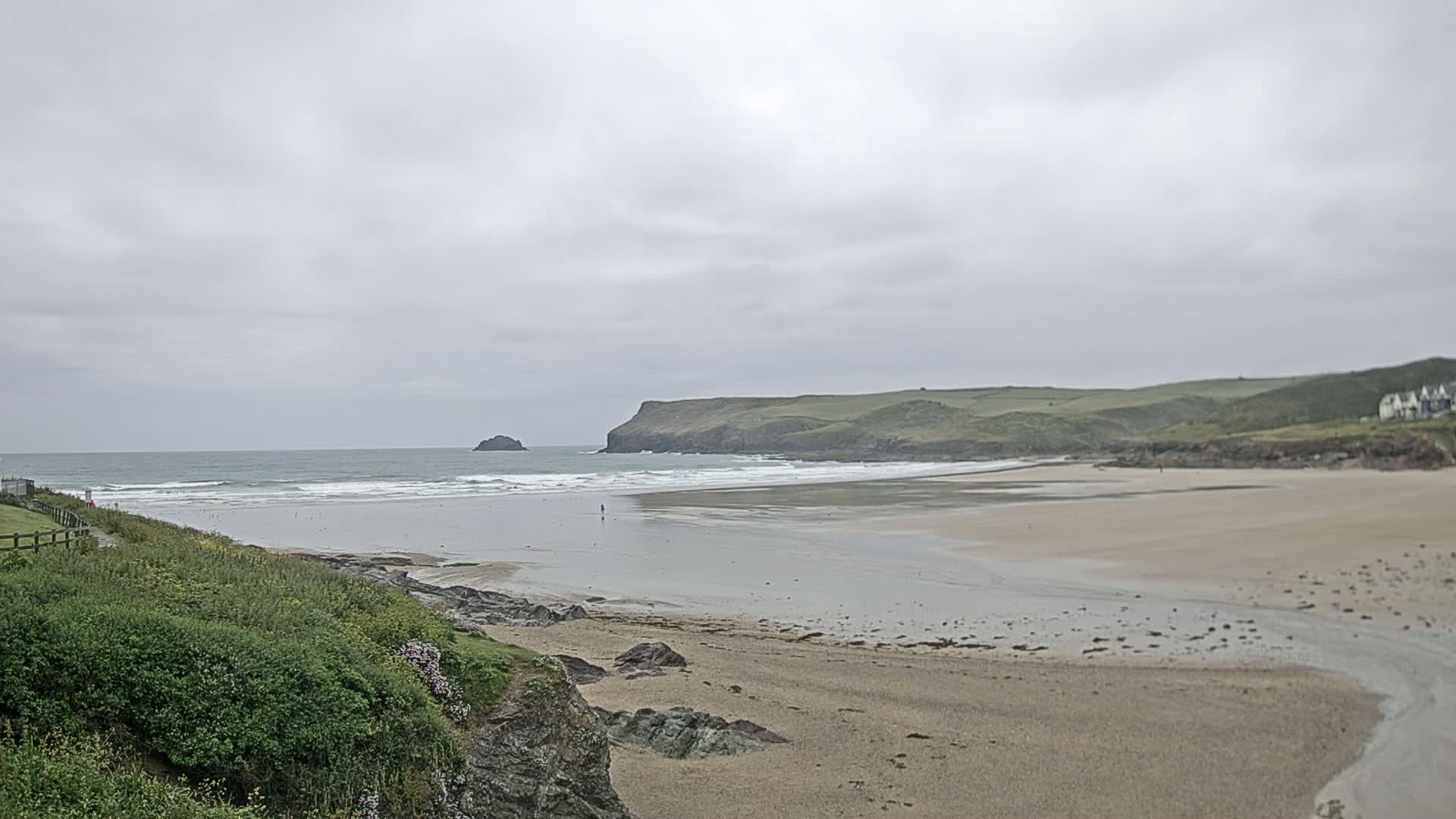 A wide sandy beach stretches out to a calm sea under a cloudy sky, with green hills and a small island in the distance.