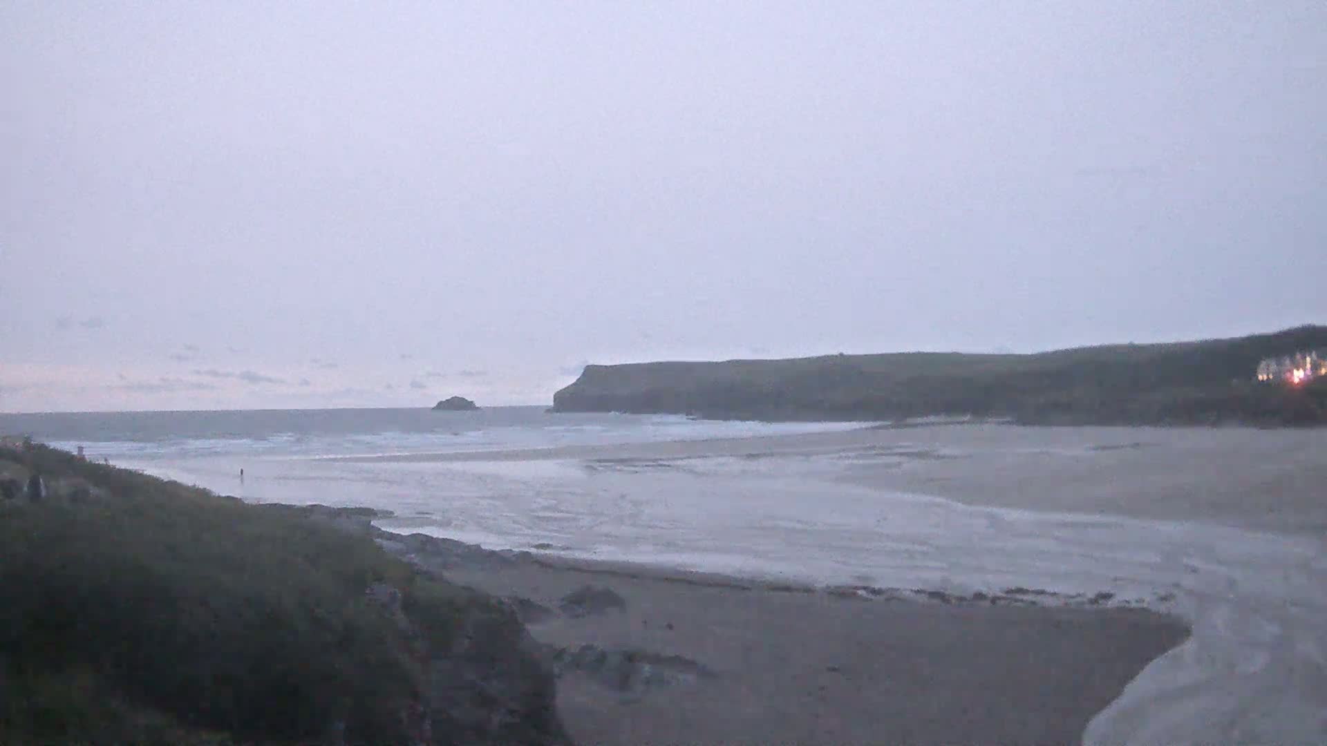 A wide, sandy beach and calm ocean are visible under an overcast sky, with a distant headland and small island in the background.