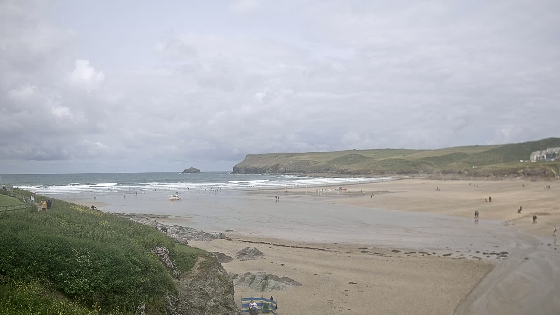 A wide sandy beach stretches out to a gently breaking ocean under a cloudy sky, with a few people scattered across the sand and in the water.