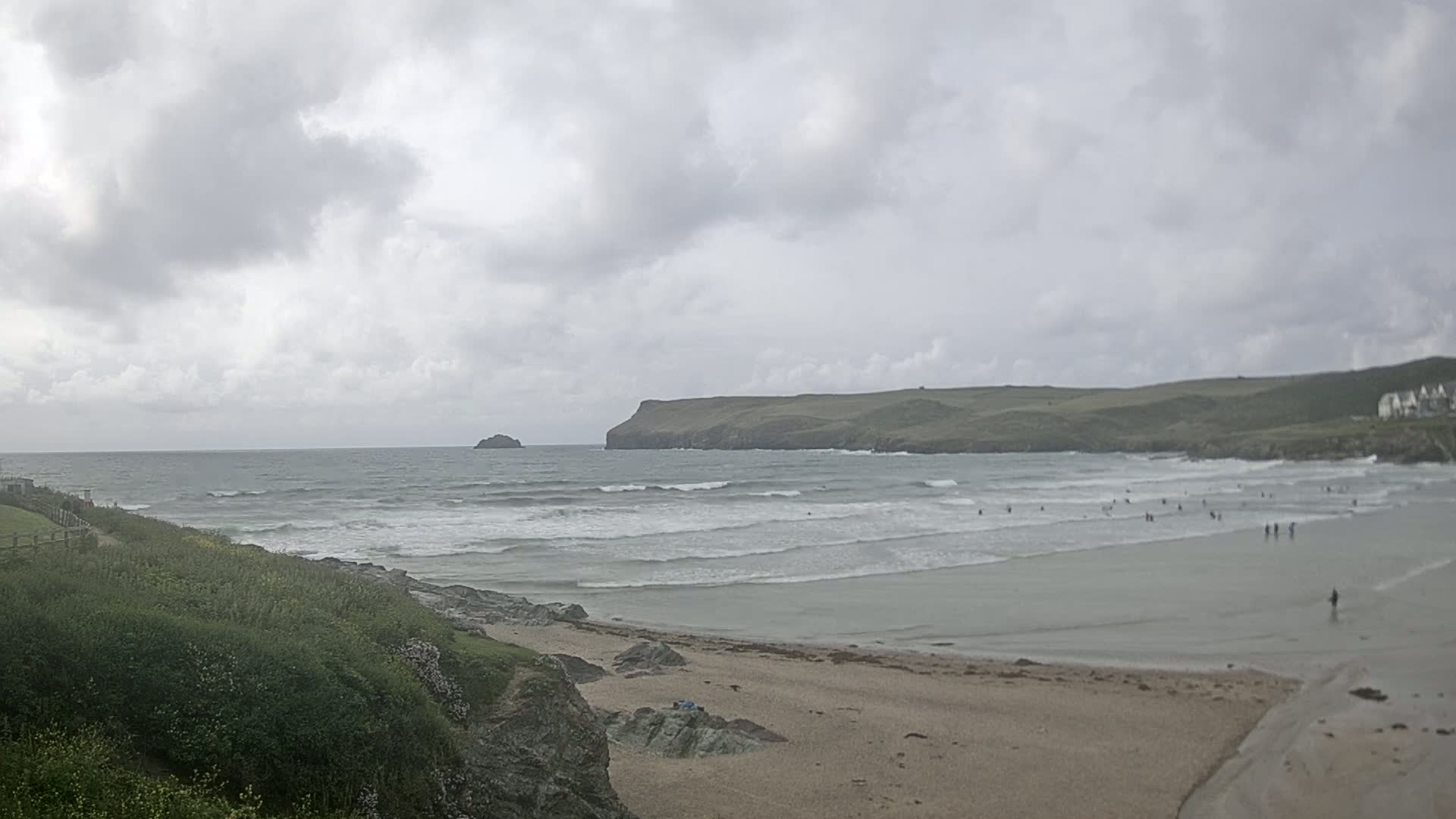 A sandy beach with small waves under an overcast sky features several people in the water and on the shore, with a rocky cliff and distant landmass visible.