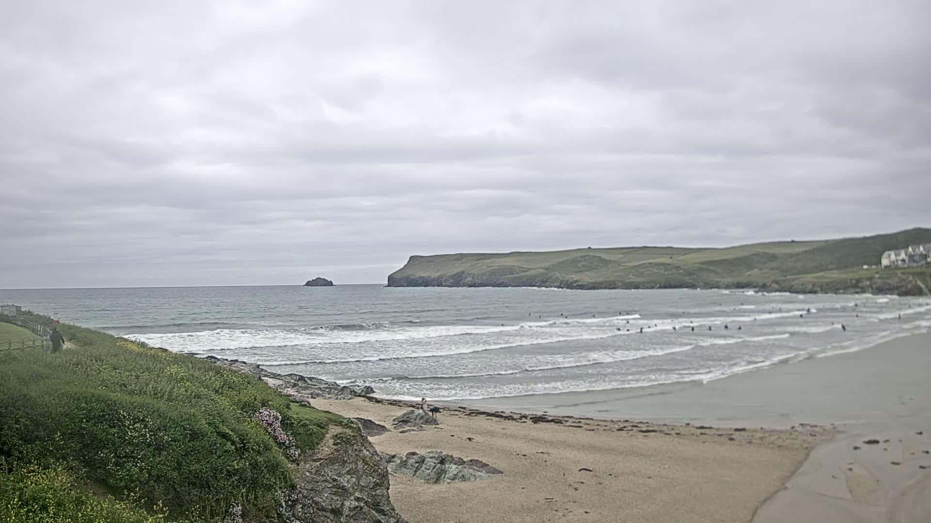 A sandy beach with several surfers in the water, under a cloudy sky, is backed by green hills and a small island in the distance.