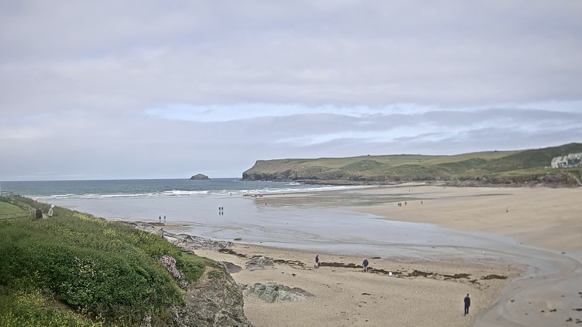 A wide sandy beach stretches out to a calm ocean under a cloudy sky, with a few people scattered along the shore and a small island visible in the distance.