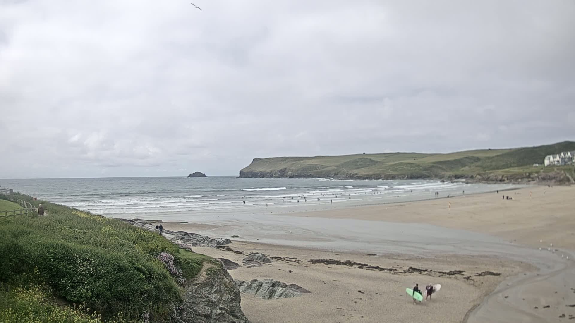 A sandy beach with people surfing and walking, backed by green hills under an overcast sky.